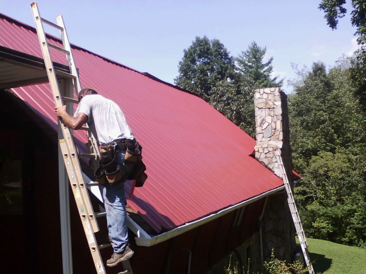 Roofer on a ladder beside a red metal roof and stone chimney — Soffit & Fascia Repair by Roofing Brothers in Etta